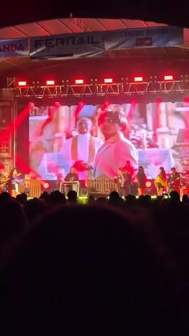 The video shows a concert stage with a large screen displaying images of people in various outfits. In front of the screen, there's a live band performing for a crowd. The stage is brightly lit with red lights, and the audience is visible in silhouette.
