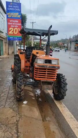 A bright orange tractor is parked on a wet, paved street next to a sidewalk. The tractor has large, knobby tires, a black seat, a steering wheel, and an open engine compartment. A dark canopy is supported by red metal poles above the driver's seat. To the left of the tractor, a tall, blue and white sign with Vietnamese text and a prominent "20%" discount is affixed to a pole. The ground around the tractor is wet and muddy, indicating recent rain. In the background, a motorcycle with a rider wearing a yellow helmet is visible on the street, and buildings line the street. The sky is overcast and appears to be cloudy.