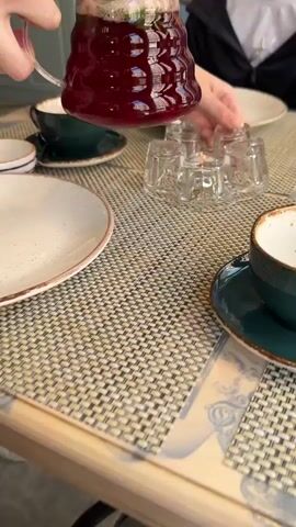 A person places a clear, beehive-shaped pitcher filled with a red beverage onto a clear, multi-faceted glass warmer on a table covered with a patterned straw mat. The table is set with plates and teacups in various patterns, some of which are speckled white with brown rims and teal saucers. A milkshake or similar beverage is visible in the background.  Someone else is sitting at the table, partially visible in a black jacket and white shirt.