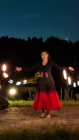 The video opens with a young woman in a long, flowing red and black skirt and a dark top, barefoot, spinning poi ignited with flames in a nighttime outdoor setting. She is positioned in the center of the frame with a dark, wooded area and a body of water in the background, illuminated by a warm-toned light orb. As the video progresses, a second woman, also in a skirt and dark top, appears on the left side of the frame, also spinning ignited poi. The two women appear to be performing together, with their movements coordinated and their flames creating streaks of light in the darkness. The camera remains focused on the performers as they continue their routine.