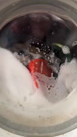 The video shows the interior of a washing machine in operation. The drum contains soapy water and several items of colorful clothing – red, blue, white, and green – tumbling around. The suds are visible on the water surface and clinging to the inside of the machine's door. The machine's drum and door appear to be made of stainless steel.