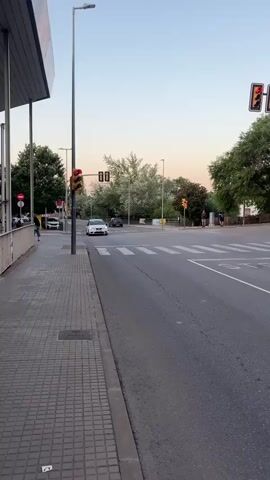 A white car is stopped at a red traffic light at an intersection. The car is facing forward, and its headlights are on. There are trees and buildings in the background, and the sky is clear and blue. Another car is visible in the distance, also stopped at a red light. A pedestrian is walking across the street in the foreground. The video is shot from a low angle, looking up at the car and the intersection. The lighting is soft and diffused, suggesting it is either early morning or late afternoon.
