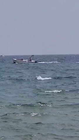 A wide shot shows a small white and red motorboat with two occupants navigating choppy, blue-green ocean water under an overcast sky. The boat is moving from left to right across the frame, leaving a wake behind it. In the distance, on the horizon, other faint shapes of boats can be seen. The foreground is dominated by the textured surface of the ocean waves. The overall scene is one of moderate sea conditions.