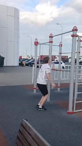 The video shows a person performing pull-ups on an outdoor gym apparatus. The person is wearing a white patterned shirt and black shorts. They are positioned on a pull-up bar, their arms extended, then bending their elbows to pull their body upwards. After completing a repetition, they lower their body back down. The background consists of a parking lot with cars and trucks, and a cloudy sky. The gym apparatus is made of metal with red caps on the poles. The ground is covered with a dark, textured surface.