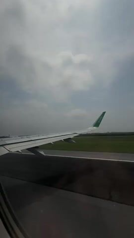 The video captures a view from an airplane window, looking out over the aircraft's wing while on an airport runway in Apodaca, Mexico. The prominent subject is the airplane's white wing, which has a distinct green wingtip featuring the white text "VIVO."

Outside the window, a dark asphalt or concrete runway stretches alongside a wide expanse of bright green grass. The sky above is cloudy, but bright, indicating it is daytime. Between the 0:02 and 0:03 mark, another airplane can be briefly seen further down the runway, suggesting active airport operations. The aircraft from which the video is shot appears to be stationary or moving slowly, possibly taxiing or waiting for departure/arrival clearance. The "VIVO" branding on the wing identifies the airline as Viva Aerobus, a Mexican carrier, which aligns with the given location of Apodaca, Mexico, home to Monterrey International Airport, a hub for the airline.