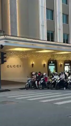The video shows a city intersection in front of a building with the Gucci logo on it. The building also has lights strung around its perimeter. People on scooters are stopped at a crosswalk, waiting for the traffic light to change. A black Mercedes SUV and a silver taxi are also waiting at the intersection. The crosswalk is marked with white stripes. The sky is overcast.