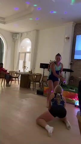 The video shows two young women dancing in a dimly lit room with colorful disco lights projected onto the ceiling and floor. The room appears to be a living area or party space, with wooden floors and modern furniture. One woman, wearing a blue and yellow athletic outfit, is standing and dancing energetically, with her arms raised and her body in a dynamic pose. The other woman, in a similar blue and yellow outfit, is kneeling on the floor in front of her, facing away from the camera, and appears to be participating in the dance or reacting to the standing woman's movements. In the background, a person is seated at a table, and other furniture and decorative elements are visible, suggesting a social gathering. The overall atmosphere is festive and lively.