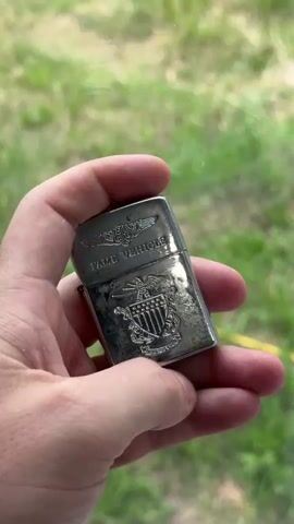 A hand holds a tarnished silver lighter. The lighter has an insignia of an eagle and shield. Above the insignia is the text "FAME VEHICLE". The person opens the lighter to show the inside. The background is blurry green grass.