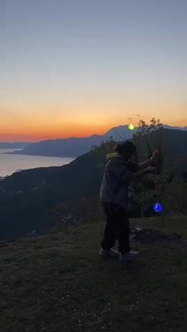 A person stands on a grassy hilltop at sunset, overlooking a large body of water (likely a bay or fjord) bordered by mountains and hills. The sky displays vibrant orange, pink, and purple hues as the sun dips below the horizon. The person, dressed in a dark jacket with a lime green/yellow lining, dark pants, light sneakers, and a dark hat, is actively spinning two glowing, spherical objects. These objects change colors, appearing as red, yellow, green, blue, and purple throughout the video, suggesting a performance with light-up poi or a similar prop. The activity takes place against the scenic backdrop of the darkening landscape, with hints of a coastal settlement visible below. The location is Bogdašići, Montenegro. No visible text is present.