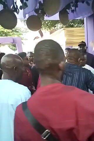 The video shows a gathering of people outdoors, seemingly at an event or celebration. People are dressed in various casual and formal attires, with some wearing traditional African clothing. The camera moves through the crowd, focusing on different individuals and small groups. There's a structure decorated with purple fabric and woven decorations. The atmosphere seems lively with music or chatter in the background. The setting appears to be a shaded outdoor area, possibly a garden or event space.