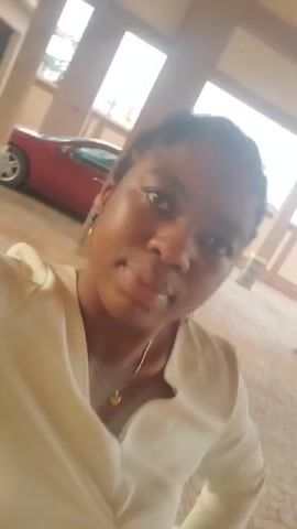 The video shows a woman with dark skin and dark hair styled in small braids. She is wearing a light-colored, button-down shirt or blouse and gold jewelry, including a ring on her left hand and a necklace. She is holding the camera at arm's length, capturing a selfie-style view of her face and upper body. The background appears to be an outdoor or semi-outdoor space with tiled flooring and supporting pillars. A red car is visible in the background. The woman is looking at the camera, and her facial expression is neutral. The camera angle shifts slightly throughout the video.