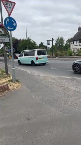 A light blue and white camper van drives through a roundabout. A dark grey sedan drives through the roundabout as well. In the background, there are buildings, trees, and a street sign. The sky is overcast.
