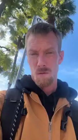 A medium-shot video presents a man with fair skin, light brown hair, and a short, reddish-brown beard. He's dressed in a black hoodie and an unzipped mustard-colored jacket. He has a black backpack with white accents. He is looking directly at the camera, with a neutral expression. The background reveals palm trees, a blue sky, and a sign with the letter “M.” The perspective indicates the camera is held at chest level.