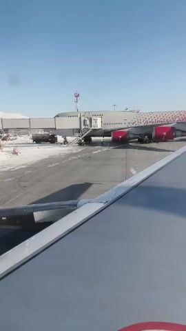 The video shows a wing of a plane on the ground, viewed from inside the aircraft. In the distance, a large red and white airplane is parked at a gate, with stairs leading to its entrance.  A person in a yellow vest can be seen near the plane. Steam or smoke rises from the ground nearby, possibly from de-icing. The sky is clear and blue. The ground around the aircraft has patches of snow.