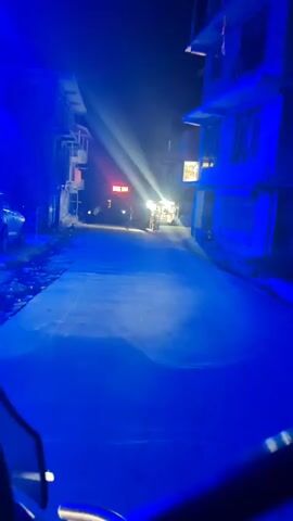 The video shows a dark street at night, illuminated by bright blue lights and a single spotlight. A person is walking away from the camera down the street towards a brightly lit stall. The stall appears to be selling various items. Buildings line both sides of the street.