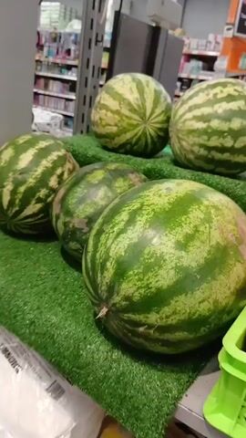 The media file shows a close-up, handheld shot of a produce display in what appears to be a supermarket or market in Bazar-Korgon, Kyrgyzstan. The foreground is dominated by a pile of large watermelons, neatly arranged on a green, artificial turf mat. Behind the watermelons, more of the same fruit is visible, some on a slightly elevated shelf. To the right, a bright green plastic crate overflows with a variety of apples, with some yellow apples also visible in another green crate behind them. The background shows shelves stocked with various goods, suggesting an indoor retail environment. There is no visible indication of the time of day or weather. The focus is on the fresh produce, and no people are visible. The overall impression is one of abundance and fresh offerings. Some text is visible on a white plastic bag in the lower left corner, but it is blurry and unreadable.