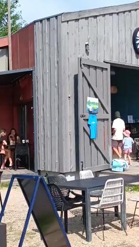 A wide shot shows the exterior of a gray, wooden building with large double doors that are open, revealing the interior of what appears to be a cafe or shop. Tables and chairs are set up outside in front of the building. A woman in a white top and skirt stands inside the doorway. A child in a pink hat and shorts walks into the building, followed by another person. A blue t-shirt hangs on the exterior of the building. To the left of the building, another section is visible with red walls and a window. Inside this section, two women are seated at a table. The ground outside is covered in gravel.