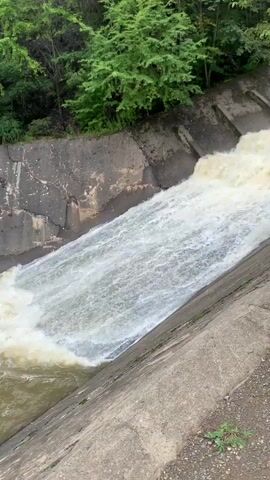 A steep concrete channel is filled with fast-flowing, turbulent water. The water appears muddy and churns white as it cascades down the channel. The sides of the channel are lined with concrete, and the channel is surrounded by lush green trees. The water level is high, suggesting recent heavy rainfall or a controlled release from a dam or reservoir. The scene is static, with the camera fixed on the flowing water.