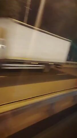 The video shows a view from inside a vehicle traveling at night. The road is visible, with lane markings illuminated by streetlights. Trees and buildings line the side of the road. There are occasional streetlights and what appear to be traffic signals. The video is blurry due to the motion of the vehicle.
