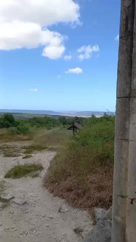 A vertical video captures a landscape under a blue sky with scattered white clouds. A dirt path with scattered rocks and dry grass leads down into a valley filled with green trees and bushes. In the distance, a body of water is visible, possibly the sea, with a faint line of land on the horizon. A small, dark-colored structure, possibly a gazebo or shelter, is partially visible among the trees. The right side of the frame shows a stone structure, possibly a wall or building facade, made of rough-hewn stones. The video is still and shows no people or animals.