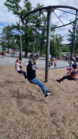 This is a video of children on a merry-go-round in a park. There are several children on the merry-go-round, some are swinging and some are sitting. The merry-go-round is on a wood chip surface. There are trees in the background and other playground equipment. The sky is blue with some clouds.