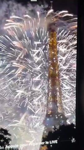 This is a vertical, first-person video of fireworks exploding around the Eiffel Tower at night. The fireworks are predominantly white and gold, with some blue sparks. The Eiffel Tower is illuminated in gold and appears to be the central focus of the display. Smoke from the fireworks drifts around the tower. There is text at the bottom of the screen that reads "Appuie pour regarder le LIVE," which translates to "Press to watch LIVE."