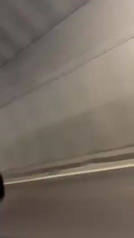 A static, close-up shot filmed from inside a car driving through a tunnel reveals a blurry view through the driver's side window. The interior of the car, specifically the driver's side mirror, is visible in the foreground. The tunnel's interior is concrete, with evenly spaced overhead lights running along its curved ceiling. The car is moving at a considerable speed, blurring the details of the tunnel walls and markings. The side mirror shows a distorted reflection of the driver. The overall impression is of a fast-moving vehicle navigating a concrete tunnel.