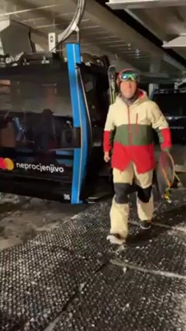 A man in ski gear, wearing a red and white helmet with blue goggles and a multi-colored jacket, is walking and smiling toward the camera. He is carrying a snowboard. He has just exited a building next to what appears to be a gondola. He walks past signs indicating the direction to the snow park. The environment is an indoor facility with artificial snow.