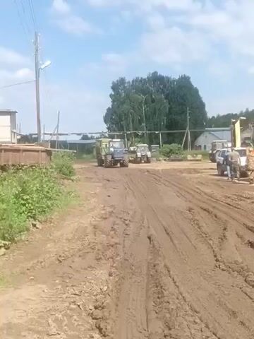 A drone flies over a muddy, rural area, showing agricultural equipment and buildings under a bright, cloudy sky. The footage begins with a view of several tractors and trailers parked on a dirt path. As the drone moves forward, it passes a brick building with a large window and then a long, wooden structure partially obscured by overgrown weeds. The scene transitions to a wide shot of a long, white building with a metal roof, suggesting it might be a barn or storage facility. The drone continues to capture the environment, revealing more of the agricultural setting.
