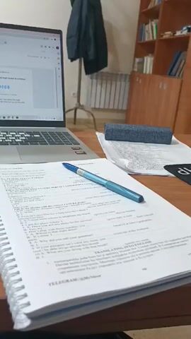 On a brown, shiny, wood desk sits a laptop, a notebook, a blue pen, and white wired earbuds. The laptop screen displays a login prompt with the code 774085. The notebook is open to a page of English grammar exercises. A blue eraser sits atop some papers, also on the desk. The background reveals a classroom setting with other wooden desks and chairs. A wooden cabinet filled with books is visible in the background, along with a coat hanging on a stand. The camera pans slightly, revealing blue denim-clad legs and other desks in the background, suggesting the scene is a student studying in a classroom.