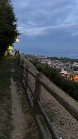 A person walks on a dirt path next to a wooden fence on a grassy hillside overlooking a town by a body of water. The sky is cloudy and the time appears to be dusk. The town has lights on and appears to be illuminated by streetlights and building lights.
