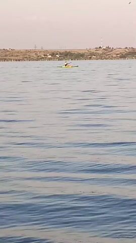 A person is kayaking on a body of water. The kayak is yellow. The person is wearing a white shirt and a red hat. They are using a paddle to propel themselves through the water. The water is blue and there are some ripples on the surface. In the background, there is a shoreline with some trees and buildings. The sky is a light blue and there are some clouds.