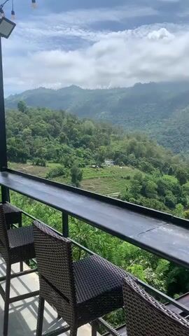 A vertical, handheld video shows a scenic view of rolling green hills and mountains under a partly cloudy sky. In the foreground, the edge of a dark metal railing and a brown wicker chair are visible, suggesting the viewpoint is from a balcony or outdoor seating area. Beyond the railing, terraced fields and scattered trees cover the slopes, with a white building visible further up one of the hills. The lighting indicates it is daytime, possibly late morning or early afternoon. The video pans slightly to the right, capturing the vastness of the landscape.