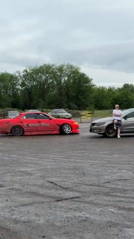 The video shows a man leaning against a gray Volkswagen CC on an asphalt parking lot with an orange Toyota Mark II parked behind it. The man is white, wearing a white t-shirt, black shorts, and a white cap, and he is leaning against the car with his right knee propped up. The Volkswagen CC is silver, and the Toyota Mark II is bright orange with decals, including "Ceramic Pro" and "Drift Taxi". The parking lot appears to be wet and there are other cars in the distance, along with trees and buildings. The sky is overcast. The Toyota Mark II does a couple of donuts in the parking lot, and then goes back to its original position.