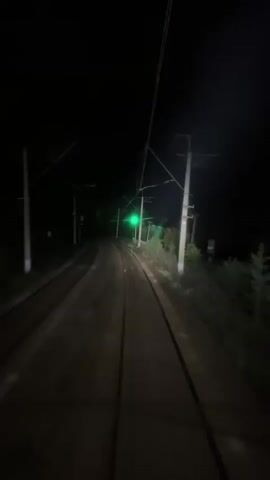 The video shows a view from a train as it travels at night. The train tracks are visible in the foreground, curving to the left. Overhead power lines stretch across the dark sky, supported by utility poles. A green railway signal is illuminated in the distance, indicating the track ahead is clear. The surrounding environment is dark, with faint outlines of trees and foliage visible along the sides of the tracks. The footage appears to be taken from the perspective of the train's operator.