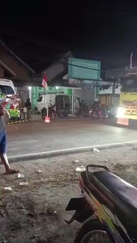 The video shows a street scene at night. In the foreground, a motorcycle is parked on the right side of the frame. In the middle ground, a man in a gray t-shirt and blue shorts stands with his arms crossed, facing a large green dump truck that is stopped on the road. The truck has its headlights on, illuminating the area. Behind the man, a white van and other vehicles are visible. To the left of the truck, a small Indonesian flag is displayed. Several other people are standing around or near the vehicles. The ground is littered with trash. The man then walks across the road, and the dump truck begins to move, passing him. The video continues to show the dump truck driving away, with its trailer visible, and another person standing on the side of the road.