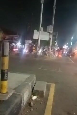 The video captures a nighttime scene at an intersection in Kedungrejo, East Java. The street is illuminated by artificial lights, including streetlights and the headlights and taillights of vehicles. Several cars and motorcycles are visible, suggesting moderate traffic. Some vehicles appear to be stopped at a traffic signal, as indicated by a red traffic light visible in the frame. The overall atmosphere is that of a typical urban intersection at night.

The scene is characterized by the blurriness typical of handheld video taken in low light conditions. The subjects are primarily vehicles, with glimpses of what might be people inside or on the motorcycles, though they are not clearly discernible. Objects like traffic signal poles and a concrete barrier are also present. The setting is clearly a road intersection, possibly in a developed area given the presence of traffic signals and overhead structures.

The activity taking place is the movement and stopping of vehicles at an intersection. The time of day is definitively night. There are no clear indications of weather, but the general ambiance is consistent with a clear night. Location cues are limited to the text on a sign, which is largely illegible due to the resolution and distance. There are no discernible emotions conveyed due to the lack of clear human subjects and the overall low-fidelity nature of the recording. The visible text on the sign is too blurry to be read.