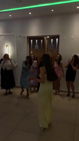 The video shows a group of women standing and clapping in a room with tiled floors. One woman wearing a long yellow dress is facing away from the camera. The other women are wearing a variety of dresses in different colors, including black, pink, blue, and floral. The room has white walls and a dark wood door in the background. The ceiling is lit with colored lights that change from green to purple to blue and then back to red.  Some of the women are holding their phones, possibly recording.