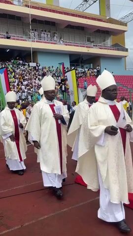 Several Catholic bishops, dressed in white chasubles with red stoles and white mitres, walk in a formal procession on a red track within an outdoor stadium in Malabo, Equatorial Guinea. They are proceeding along a red carpet laid out on the track. The stadium stands are filled with a large crowd of spectators, many dressed in white, green, and yellow, observing the event. Flags of Equatorial Guinea and Vatican City are visible in the background. The sky is overcast, suggesting daytime with cloudy weather. The scene depicts a significant public religious ceremony.