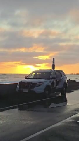 A police SUV is parked on a wet road near the ocean. The sun is setting, casting a warm glow on the clouds. Waves crash against a seawall, sending spray into the air. A pier is visible in the distance.