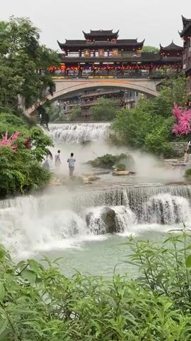 The video shows a picturesque scene in China, featuring a tiered waterfall cascading down towards the viewer, surrounded by lush greenery and various types of vegetation. In the middle ground, two people stand on a rocky platform taking pictures or admiring the view. Above the waterfall, a bridge arches across the water, supporting an ornate traditional Chinese building, likely a restaurant or teahouse, adorned with lanterns. Several people are walking on the stone steps and bridges adjacent to the building. The weather appears overcast, but the scene is vibrant and colorful. The style of the architecture, along with the presence of the waterfall and the surrounding landscape, suggests a popular tourist destination.