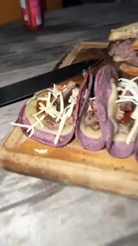 Here is a detailed description of the video.

The video shows a wooden cutting board with tacos and sandwiches on it. The tacos have purple shells and are filled with cheese and meat. The sandwiches appear to be burger patties between slices of bread. A person's hand is seen putting the top piece of bread on a sandwich. The scene is dimly lit, possibly outdoors at night. A black knife is on the board, along with a bottle of hot sauce. A water bottle is visible in the background.