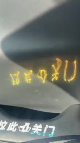 A video shows a close-up shot of a dashboard in a car, possibly from the driver's or passenger's perspective. The dashboard has what appears to be a warning light illuminated in yellow. The warning symbols are not easily recognizable, but they appear to be related to vehicle functions. Below the illuminated symbols, there's a sticker with Chinese characters. The dashboard itself is gray, and there is some sunlight shining across it, casting shadows.