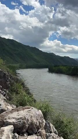 The camera films a wide river flowing between green mountains on a partly cloudy day. The river water is a muted, silty color. In the foreground, there are grey rocks and low-lying green plants. The sky is blue with fluffy white clouds and dark grey clouds. The mountains on either side of the river are covered in trees. In the distance, more mountains are visible. There is no one to be seen in the video.