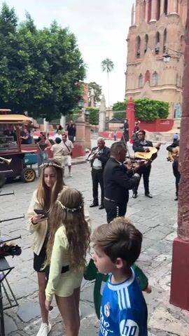 The video shows a bustling square in San Miguel de Allende, Mexico. In the background stands the Parroquia de San Miguel Arcángel, a distinctive neo-Gothic church with tall spires. A mariachi band is performing in the middle of the square, clad in traditional black suits with silver accents. Several individuals are milling about, including two young girls wearing floral headbands, and two boys in sports jerseys. One boy wears a green jersey with the name "Elias Miche" and the number "4" on the back, while the other is in a blue Real Madrid jersey. The camera is positioned from a slightly low angle, possibly from someone standing near a building's red wall. A food cart can be seen to the left, near some people sitting. The weather appears pleasant with a partly cloudy sky.