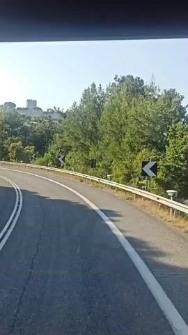 The video shows a view from inside a vehicle, likely a bus, as it travels along a paved road. The road curves to the left, with a white line marking the center and a yellow line on the right shoulder. A metal guardrail runs along the right side of the road, separating it from a grassy embankment. Trees and foliage line the embankment, with some taller trees visible in the background. A castle or fortified structure is visible on a hilltop in the distance, partially obscured by trees. Road signs with black arrows on yellow backgrounds are mounted on poles along the right side of the road, indicating curves. The sky is clear and blue, suggesting a sunny day. The interior of the vehicle is visible at the top of the frame, with a dark, possibly tinted, window or roof.
