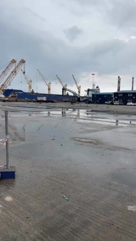 The video depicts a wet concrete area with puddles reflecting the sky and surrounding structures. In the background, a large blue ship is docked, with several cranes positioned alongside it, presumably for loading or unloading cargo. To the right of the ship, there's a blue building and several vehicles parked nearby. The sky is overcast with gray clouds. A metal pole with a blue base and warning tape is visible in the foreground on the left.