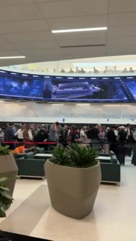 The camera pans slowly in an indoor space to reveal a crowd of people standing in line. Many have luggage, suggesting an airport or travel hub. An enormous curved screen above the crowd displays footage of a space shuttle. There are a few people sitting on lounge chairs and some potted plants nearby. Some people are interacting with each other, others appear to be waiting patiently in the queue. The overall atmosphere seems calm and orderly.
