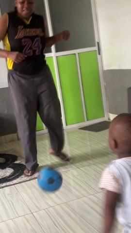 A toddler attempts to kick a small blue soccer ball inside of a house. The toddler is wearing a white outfit and a diaper. A woman in a Lakers #24 basketball jersey and grey pants stands nearby, observing the child. The toddler falls while kicking the ball and ends up on their hands and knees. The toddler then plays with the ball while calling out "Mommy." The scene is set in a living area with light-colored tile flooring. A black coffee table with stacked papers on it, a patterned rug, and a partially visible TV are in the background. A green wall element is also visible.