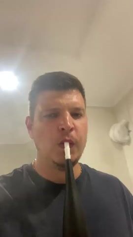 This media file features a close-up, first-person perspective video of a man in what appears to be an indoor setting. The man, with short brown hair and a light beard, is wearing a dark t-shirt and a gold chain. He is holding a black device with a white mouthpiece to his lips and exhaling a visible plume of smoke. The background is a neutral-colored wall, possibly painted or textured, with some indistinct objects or fixtures visible. There is a bright light source in the upper left corner of the frame, suggesting artificial lighting. The man's facial expression is neutral, and the overall atmosphere is casual and personal. The video captures the act of smoking, likely from a shisha pipe or vaporizer, with the smoke being a prominent element. No visible text is present in the media. The context provided indicates the location is Yablunivka, Ukraine.